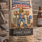 Coffee bag labeled 'Foundation of Freedom Cowboy Blend' with a graphic of a man lifting weights, placed on a rustic surface with beer bottles.