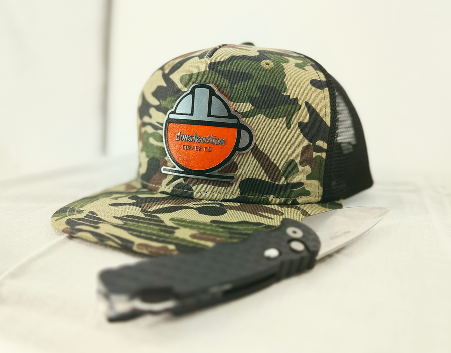 A camouflage flat bill cap with a black mesh back featuring an orange and white 'Hard Hat Mug' logo patch on the front.