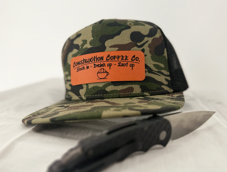 A camouflaged snapback hat with a leather patch that has text on it, sitting next to a knife.