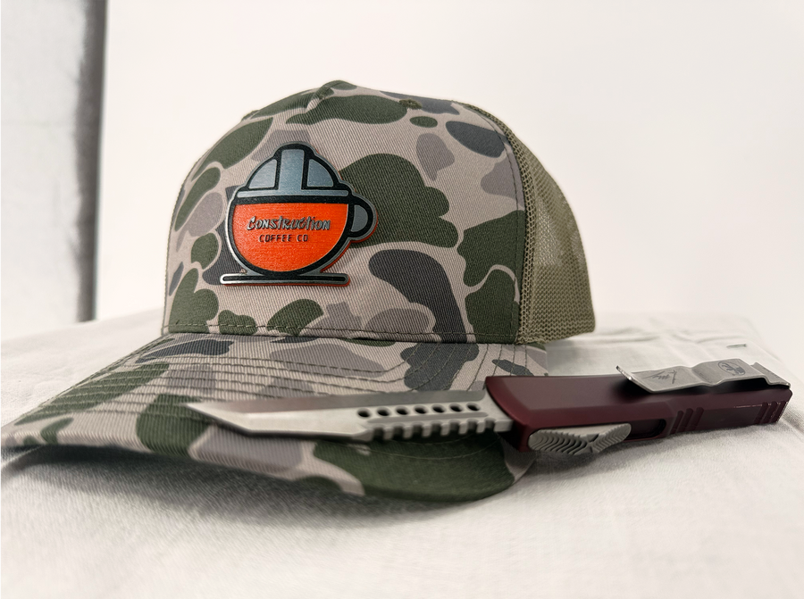 A camouflage cap with a black mesh back and an orange logo on the front, displaying a hard hat shape with the text 'HARD HAT CAFE'.