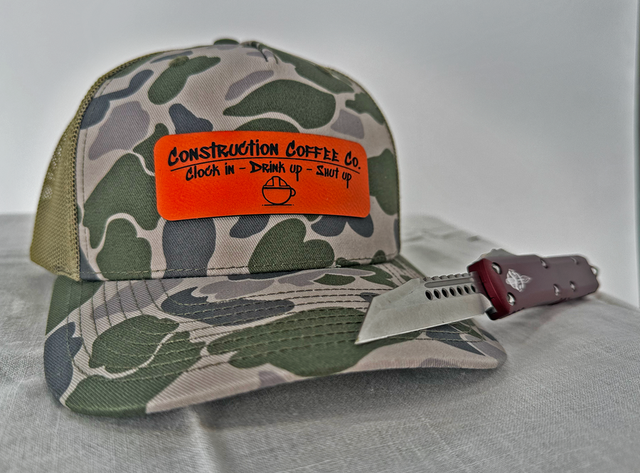 A camouflage cap with an orange high visibility leather patch on the front and a black mesh back, accompanied by a pair of sunglasses.
